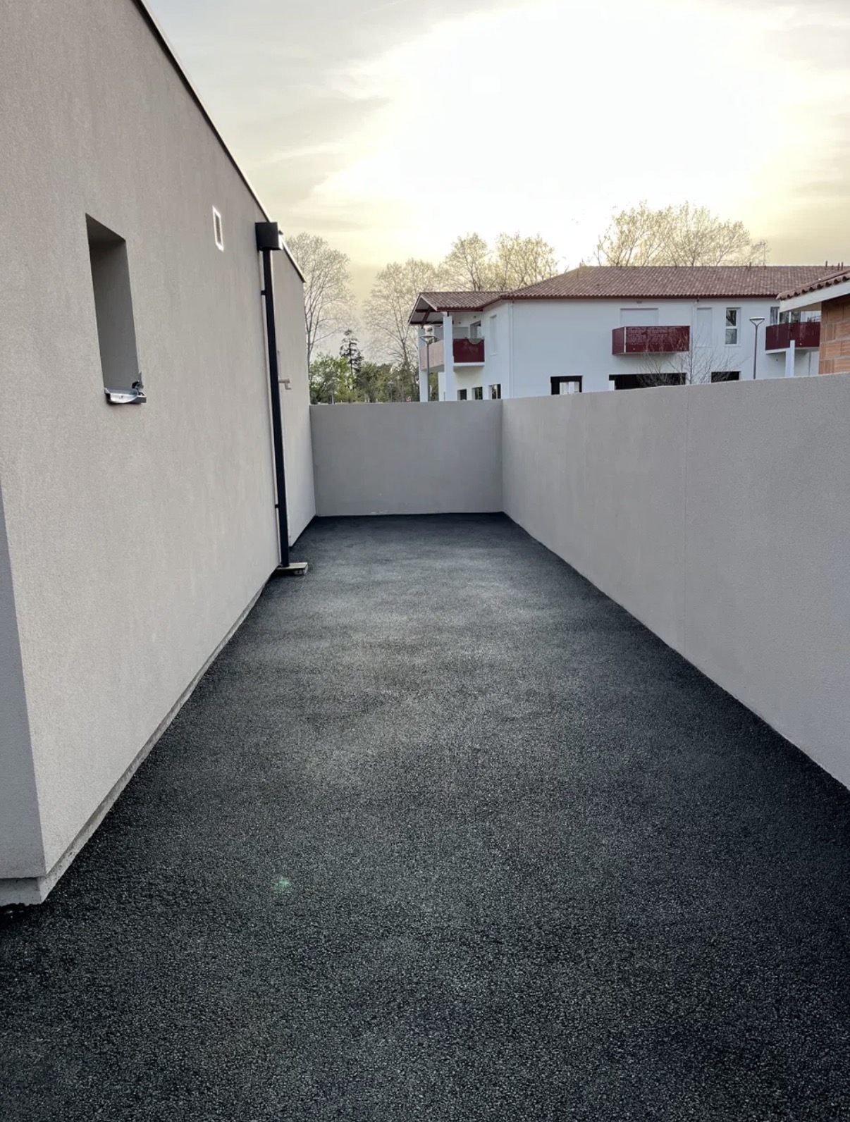 Cour close en enrobé noir fraîchement posé entre deux murs enduits clairs, finition lisse jusqu'aux rives